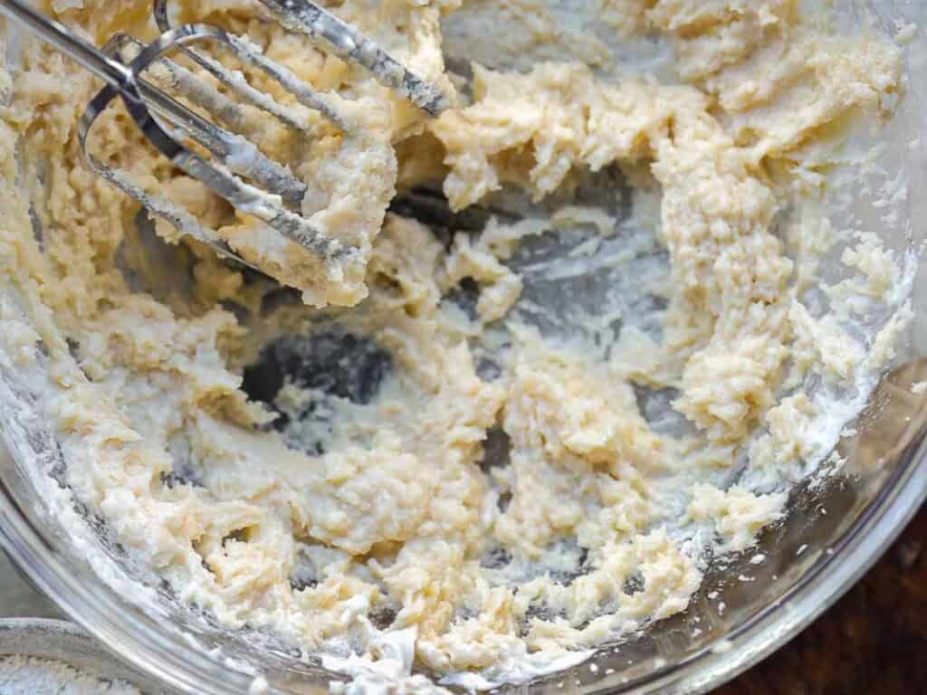 A close-up of a glass mixing bowl with creamy batter being blended by a metal hand mixer, with some flour visible on the edge of the bowl.